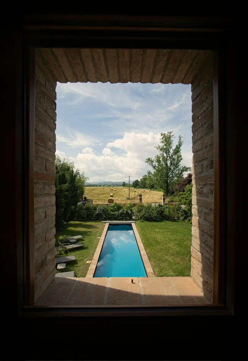 La piscina lunga nel prato vista da una finestra nel muro di pietra, con la campagna sullo sfondo.