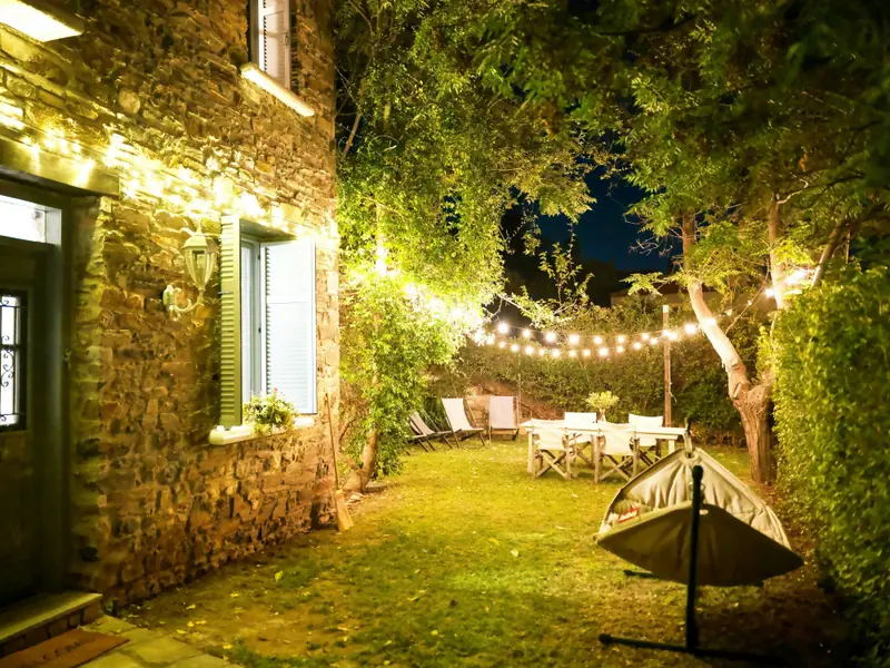 Il giardino di sera accanto al muro di pietra: luci appese tra gli alberi, la tavola apparecchiata sul prato.