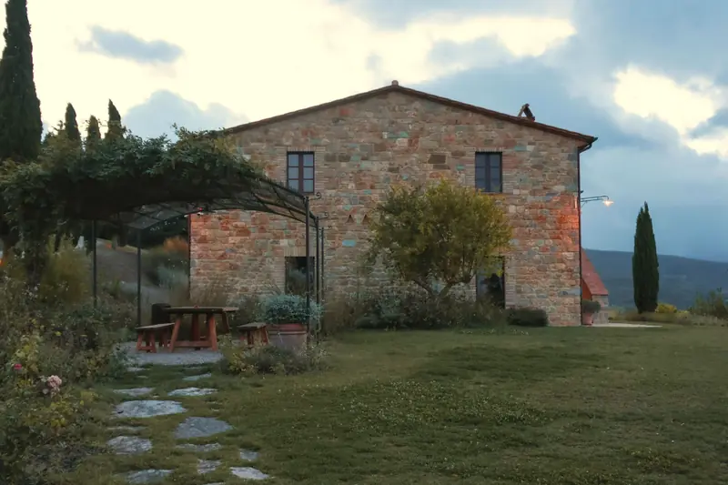 La facciata in pietra della casa, il pergolato di ferro con il tavolo di legno e il sentiero di lastre nel prato.
