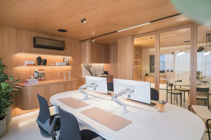 Scrivania curva bianca con due schermi, sedie nere e mensole in rovere illuminate dal basso; soffitto in legno.