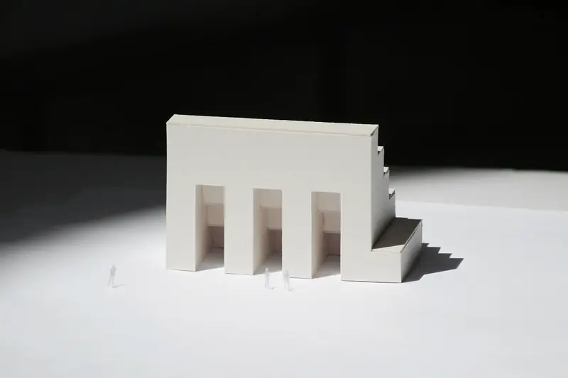 Plastico in cartoncino bianco di una casa con tre portali, illuminato di taglio su un tavolo; accanto, due figurine in scala.