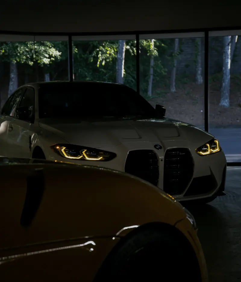 Il salone in penombra: una BMW bianca con i fari accesi dietro il cofano giallo di un'altra auto, e le vetrate sul bosco.