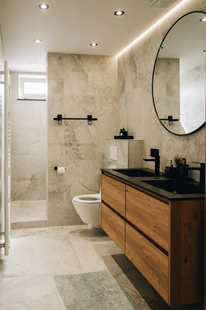 Bagno finito: rivestimento in gres effetto pietra, doccia a filo pavimento, mobile in rovere con doppio lavabo nero e specchio tondo.