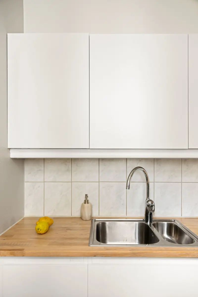Angolo cucina con pensili bianchi, rivestimento in piastrelle chiare, top in rovere massello e lavello in acciaio a due vasche.