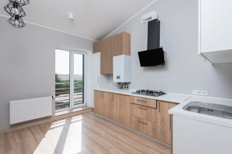 Cucina lineare con basi in legno chiaro e top bianco sotto il soffitto inclinato, portafinestra sul balcone e caldaia a parete.