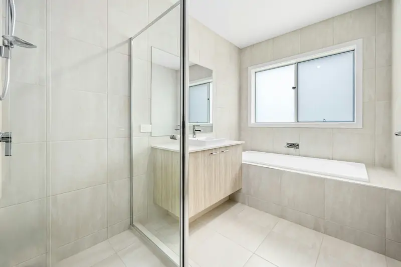 Bagno rifinito in gres chiaro: doccia con parete in vetro, mobile lavabo in legno chiaro, vasca incassata sotto la finestra.