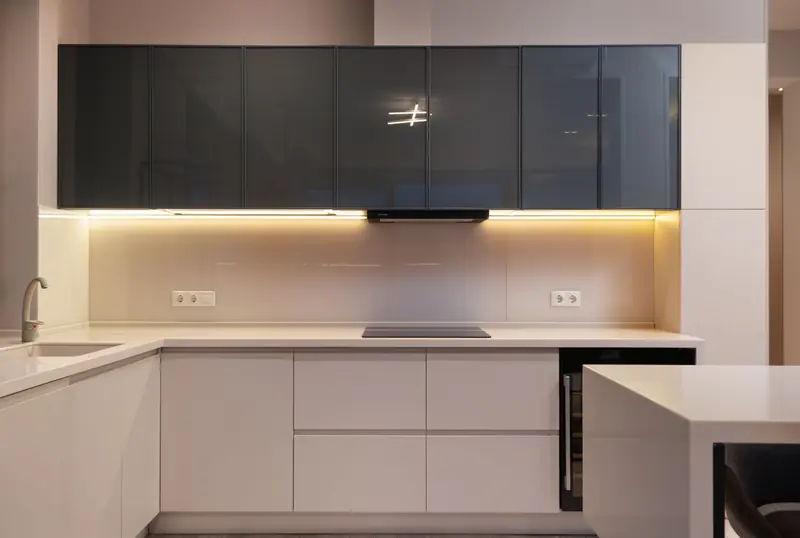 Cucina ad angolo con basi bianche, pensili in vetro scuro e luce sotto i pensili.