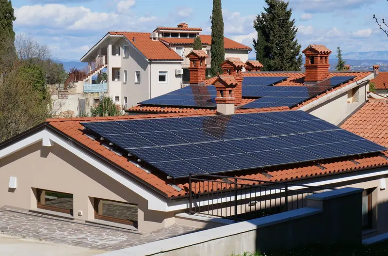 Tetto di tegole rosse coperto di pannelli fotovoltaici scuri, tra comignoli in mattoni e cipressi.