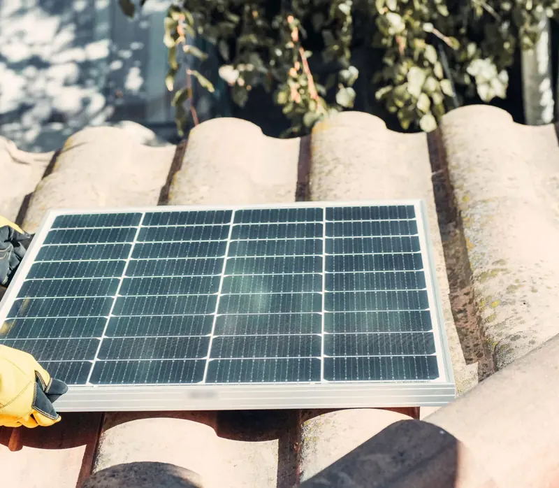 Due mani con i guanti da lavoro appoggiano un pannello fotovoltaico sulle tegole di un tetto.