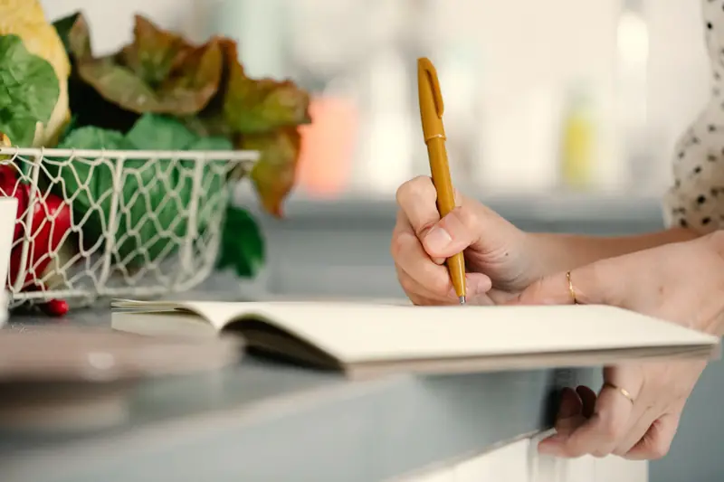 Una mano prende appunti con una penna color senape su un quaderno aperto, accanto a un cestino di verdure.
