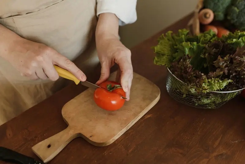Mani con un grembiule di lino tagliano un pomodoro su un tagliere, accanto a una ciotola di insalata.