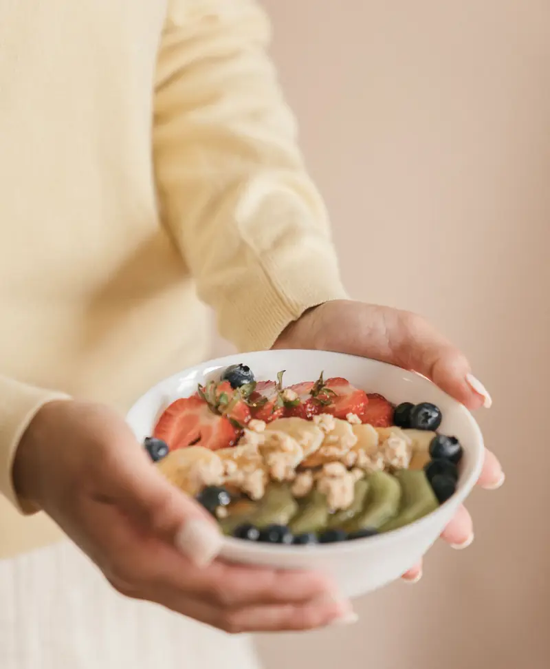 Le mani della dottoressa, con un maglione color burro, tengono una ciotola di frutta fresca, fiocchi d'avena e mirtilli.