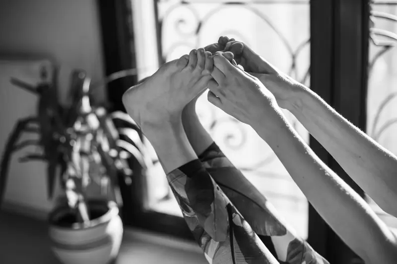 Mani che afferrano i piedi in un allungamento, accanto a una finestra.